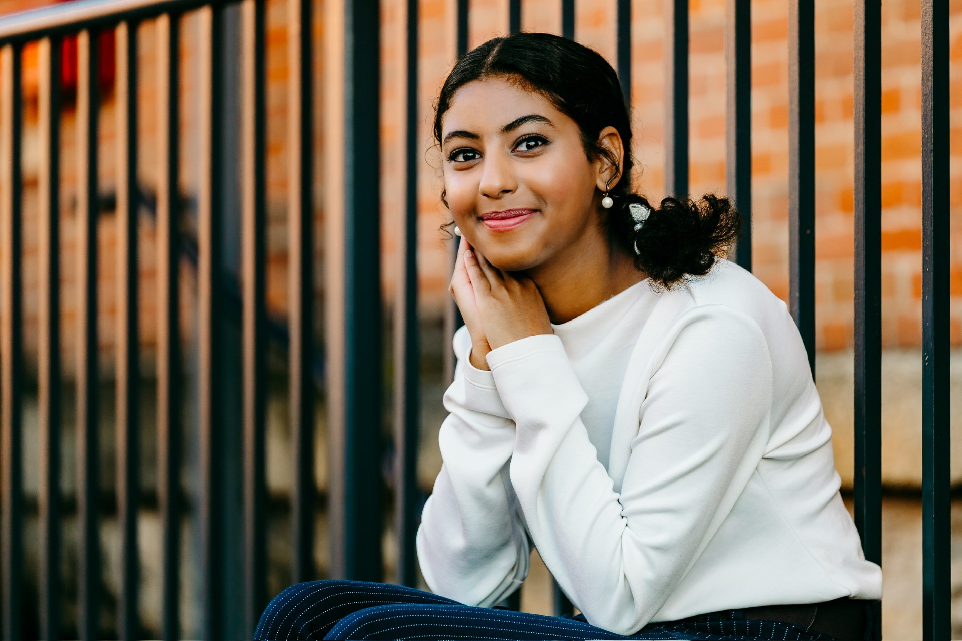 A young woman sits outdoors in front of a black metal fence, smiling gently toward the camera. She rests her chin on her hands, which are folded together, giving a relaxed and friendly expression. She has dark curly hair pulled back into a low ponytail and is wearing small pearl earrings, a light-colored long-sleeve top, and dark pants with subtle pinstripes. Behind her, a softly blurred brick wall and the vertical bars of the fence create a patterned background.