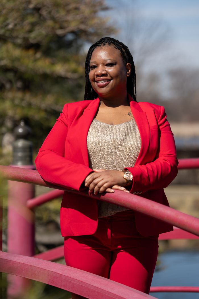 A woman stands outdoors on a small bridge, smiling warmly. She is wearing a bright red suit with a matching blazer and pants, paired with a light-colored top underneath. Her hair is styled in neat braids, and she rests her arms casually on the red railing. Sunlight highlights her face, and trees with soft greenery blur in the background, creating a calm, natural setting.