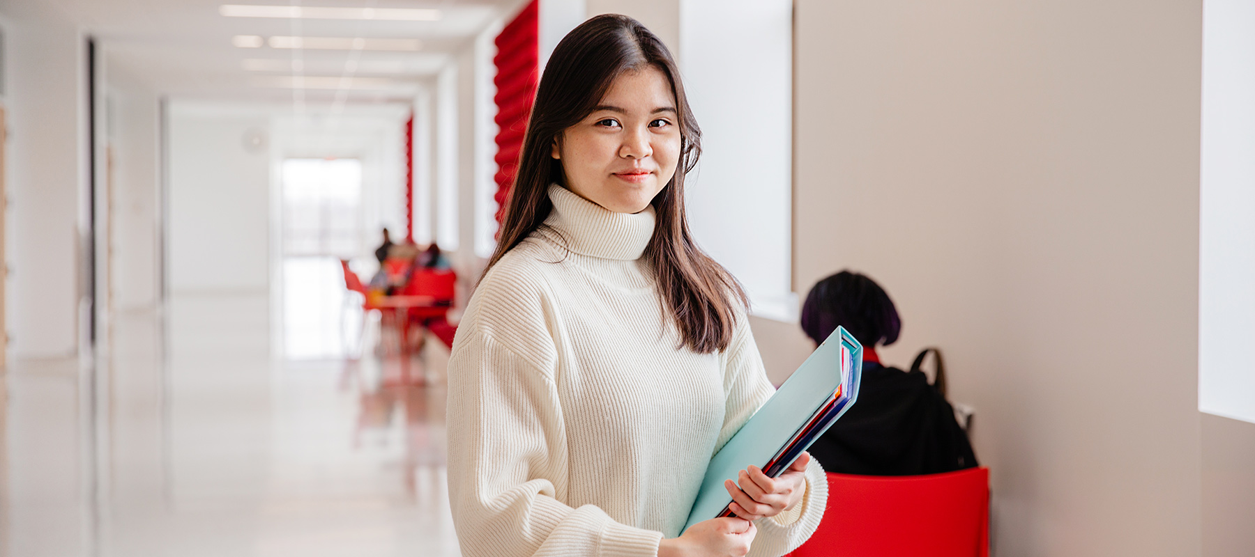 A Normandale Student in a cream sweater looks at the camera holding binders and school materials. 