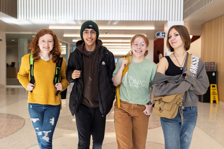 Four college students—three women and one man—smile together in a bright hallway, wearing casual clothes and backpacks.