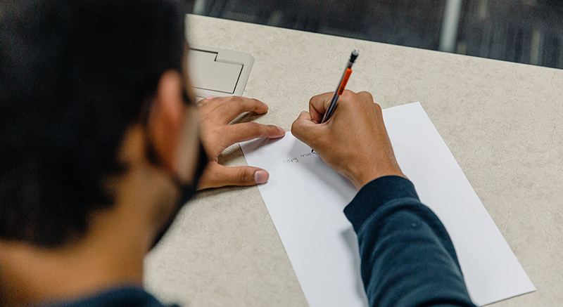 A student wearing a blue shirt writes on a white piece of paper.